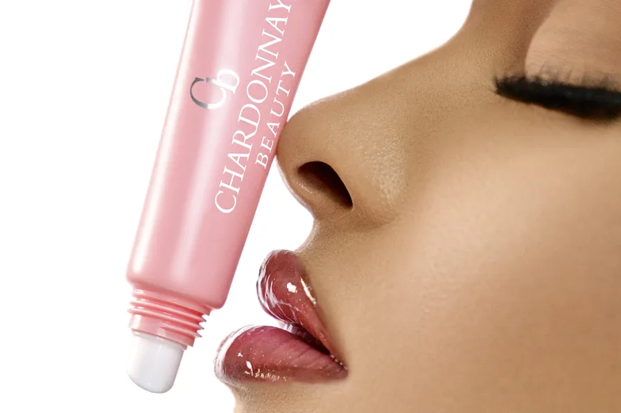A pink Chardonnay Beauty gloss tube held beside a model's high-shine rose lips, in profile against a white ground
