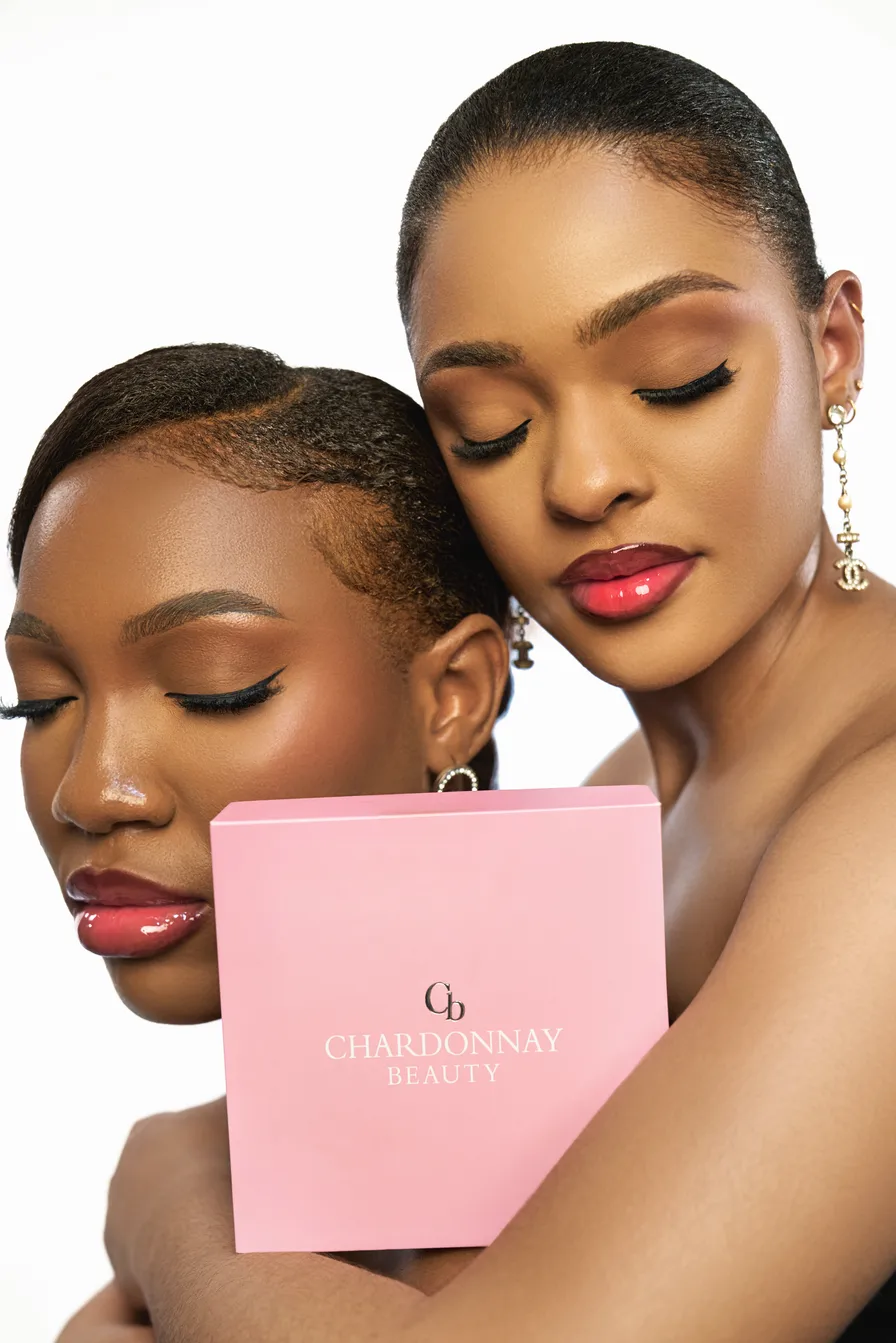 Two models resting cheek to cheek with glossy rose lips, holding a pink Chardonnay Beauty gift box