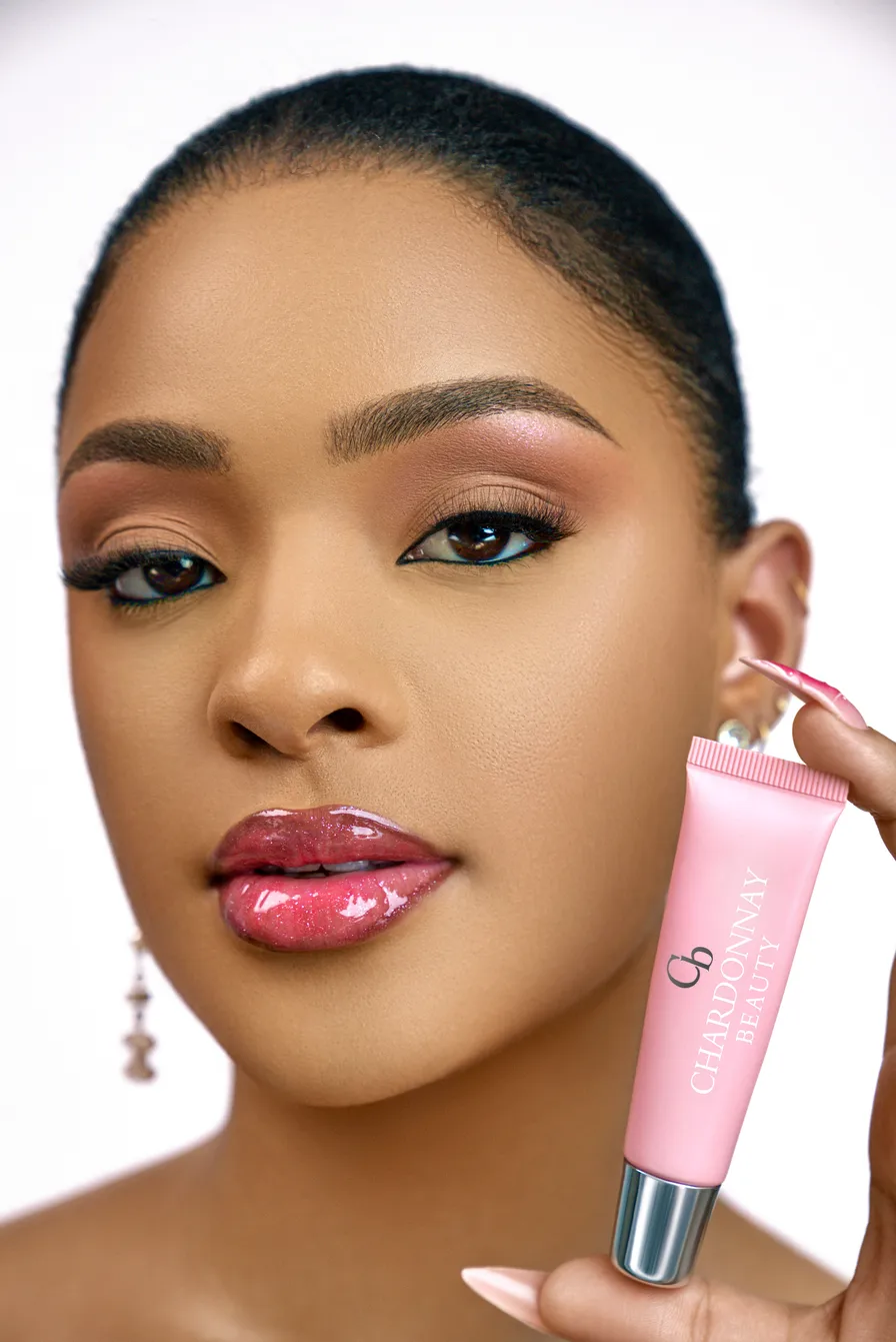 A model holding a pink Chardonnay Beauty tube beside her face, wearing a shimmering pink gloss